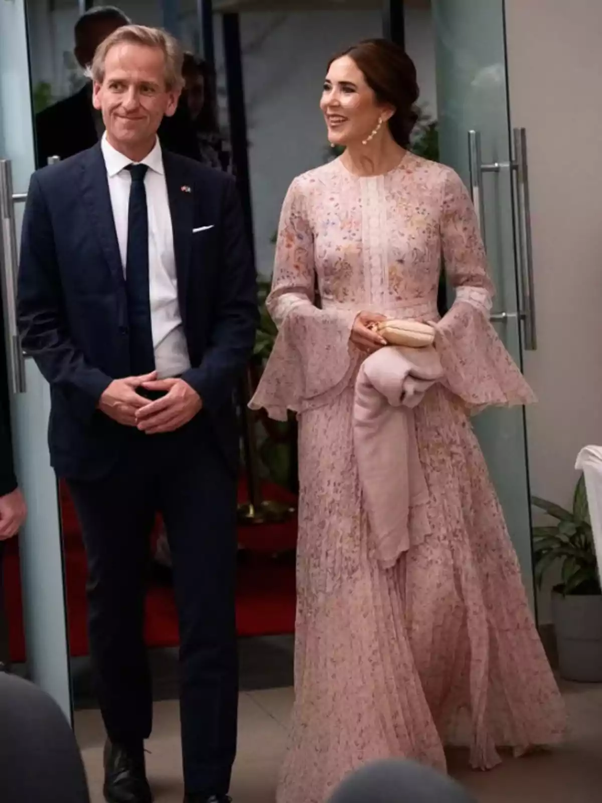 Un hombre con traje oscuro y corbata junto a una mujer con un vestido largo rosa claro y detalles florales, ambos sonríen mientras caminan en un ambiente elegante. Un hombre con traje oscuro y corbata junto a una mujer con un vestido largo rosa claro y detalles florales, ambos sonríen mientras caminan en un ambiente elegante.