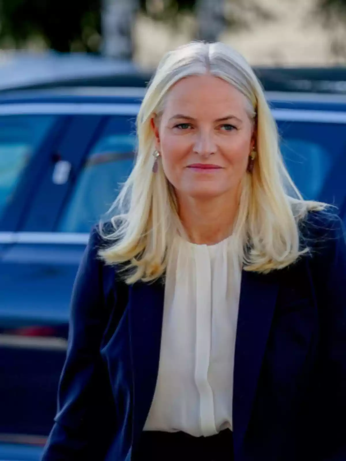Blonde woman with loose hair wearing a white blouse and dark blue blazer standing in front of a car Blonde woman with loose hair wearing a white blouse and dark blue blazer standing in front of a car