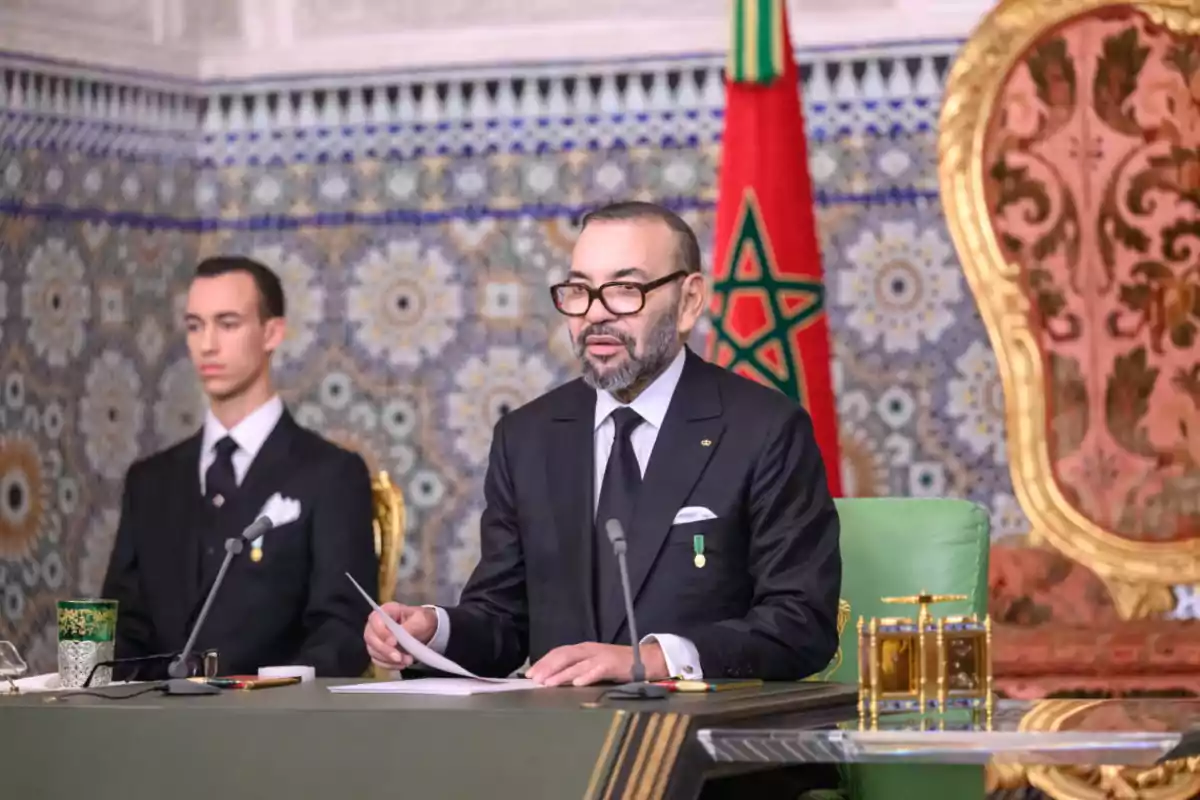 Dos hombres elegantemente vestidos con trajes oscuros están sentados en una sala decorada con mosaicos y una bandera de Marruecos, uno de ellos habla frente a micrófonos y sostiene papeles. Dos hombres elegantemente vestidos con trajes oscuros están sentados en una sala decorada con mosaicos y una bandera de Marruecos, uno de ellos habla frente a micrófonos y sostiene papeles.