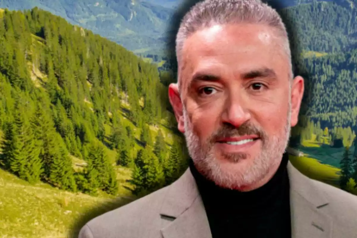 Kiko Hernández con saco gris y suéter negro posando frente a un paisaje de montañas y árboles verdes