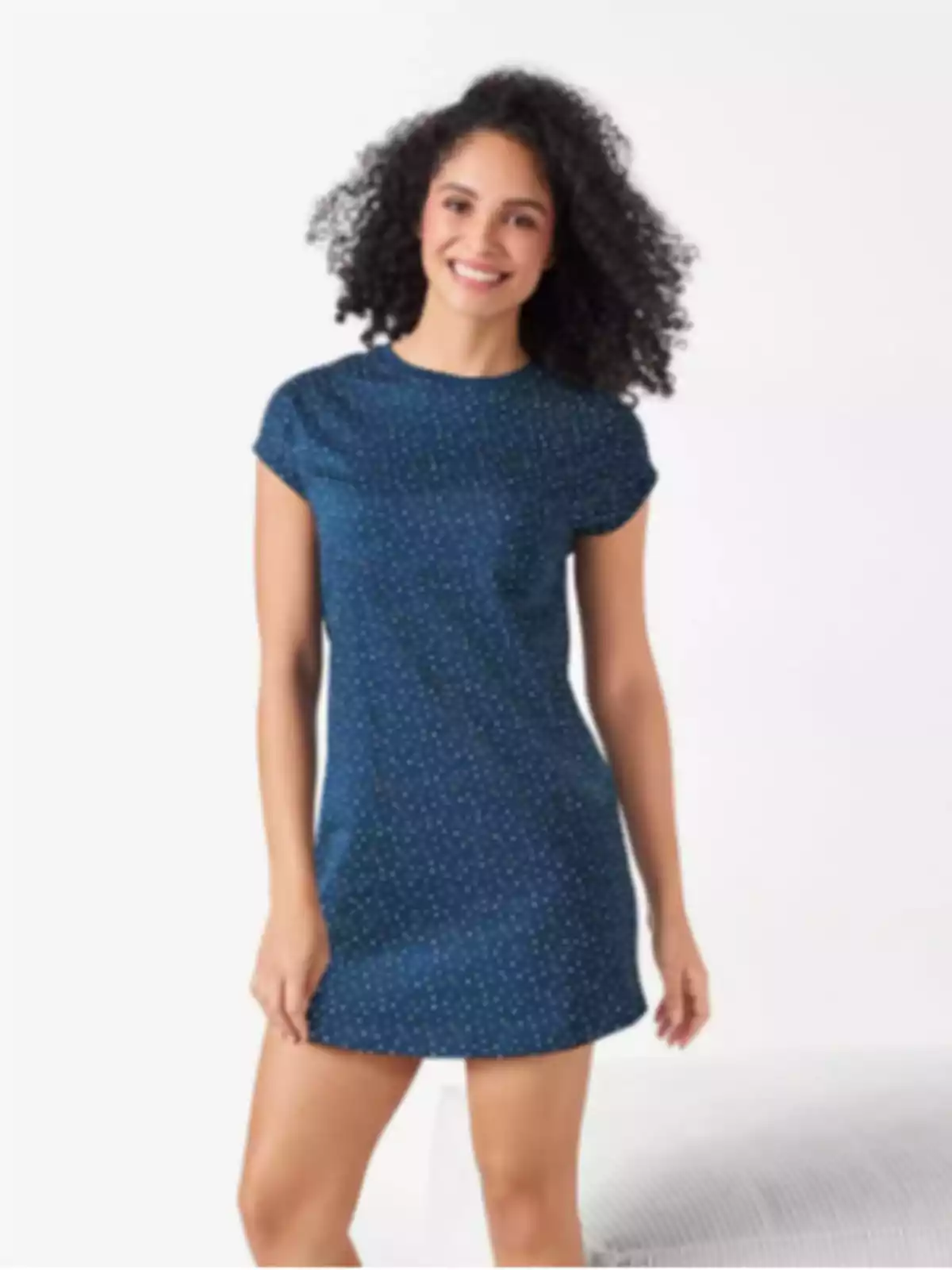 Mujer joven con cabello rizado usando un vestido azul corto con pequeños puntos blancos y sonriendo frente a un fondo claro Mujer joven con cabello rizado usando un vestido azul corto con pequeños puntos blancos y sonriendo frente a un fondo claro