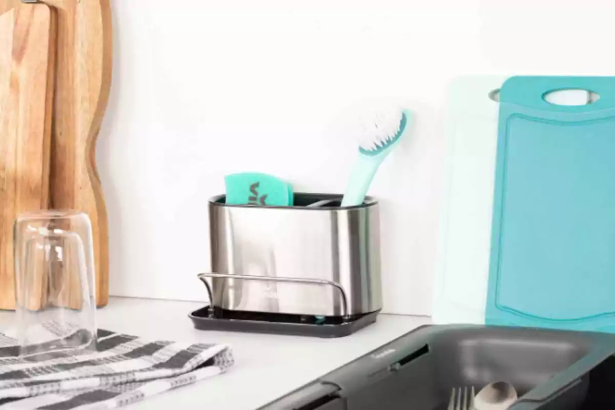 Utensilios de cocina organizados sobre una encimera blanca con tablas de cortar, vaso de vidrio, esponja, cepillo y un escurridor de cubiertos. Utensilios de cocina organizados sobre una encimera blanca con tablas de cortar, vaso de vidrio, esponja, cepillo y un escurridor de cubiertos.