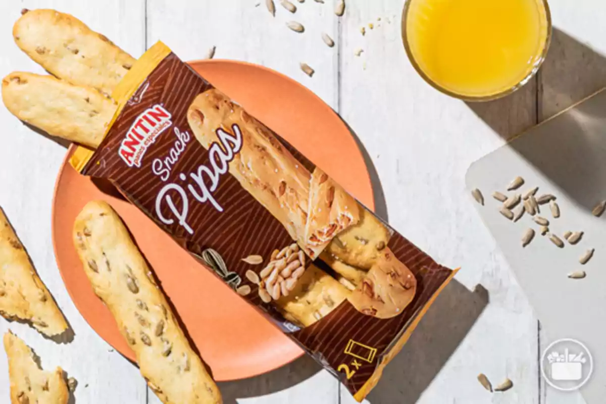 Paquete de snack de pipas Anitin sobre un plato naranja acompañado de palitos de pan, semillas de girasol y un vaso de zumo de naranja Paquete de snack de pipas Anitin sobre un plato naranja acompañado de palitos de pan, semillas de girasol y un vaso de zumo de naranja