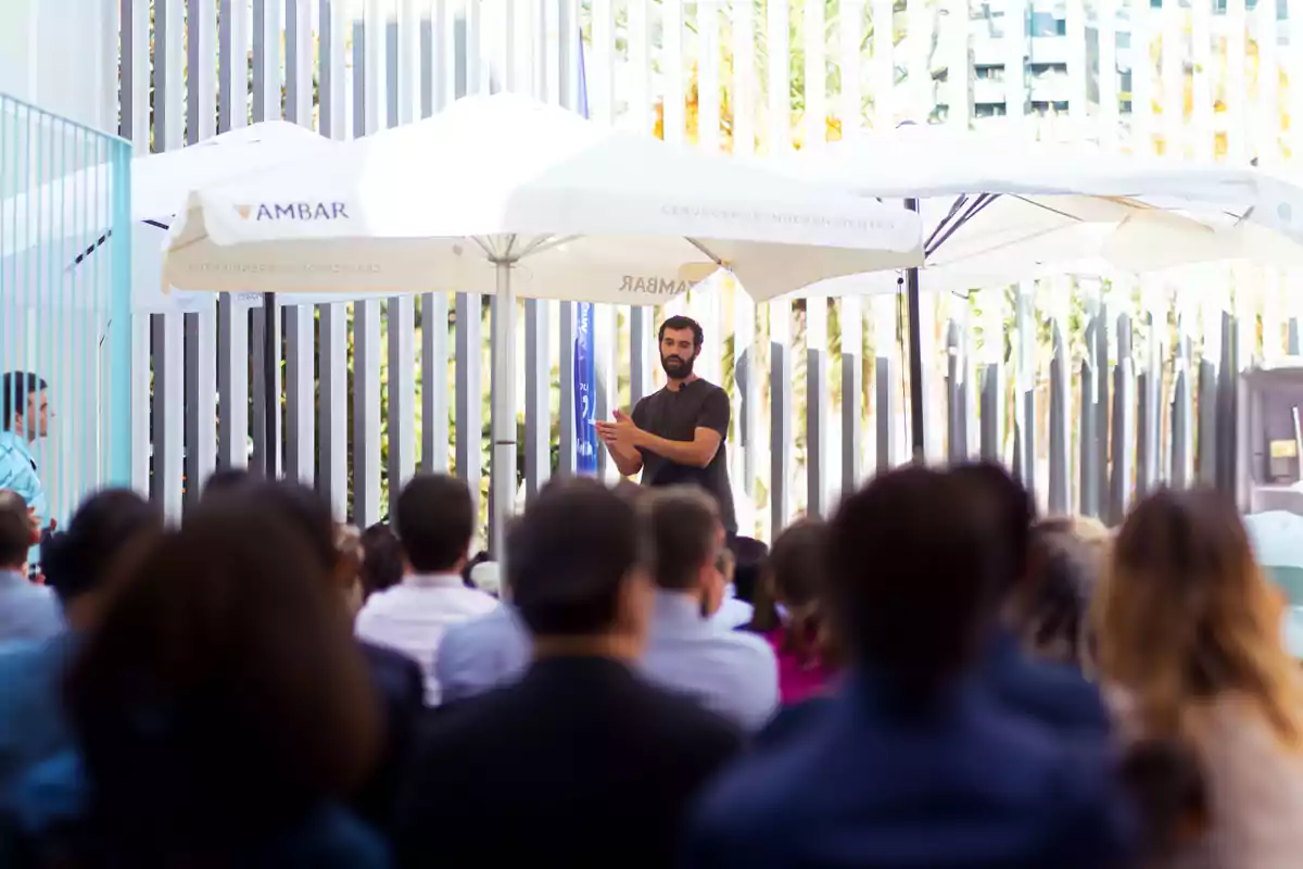 Un hombre de pie da una charla al aire libre frente a un grupo de personas sentadas bajo sombrillas blancas