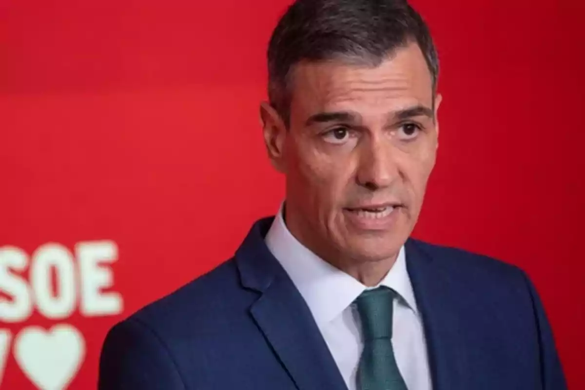 Hombre de cabello corto y canoso con traje azul y corbata verde hablando frente a un fondo rojo con letras blancas desenfocadas Hombre de cabello corto y canoso con traje azul y corbata verde hablando frente a un fondo rojo con letras blancas desenfocadas