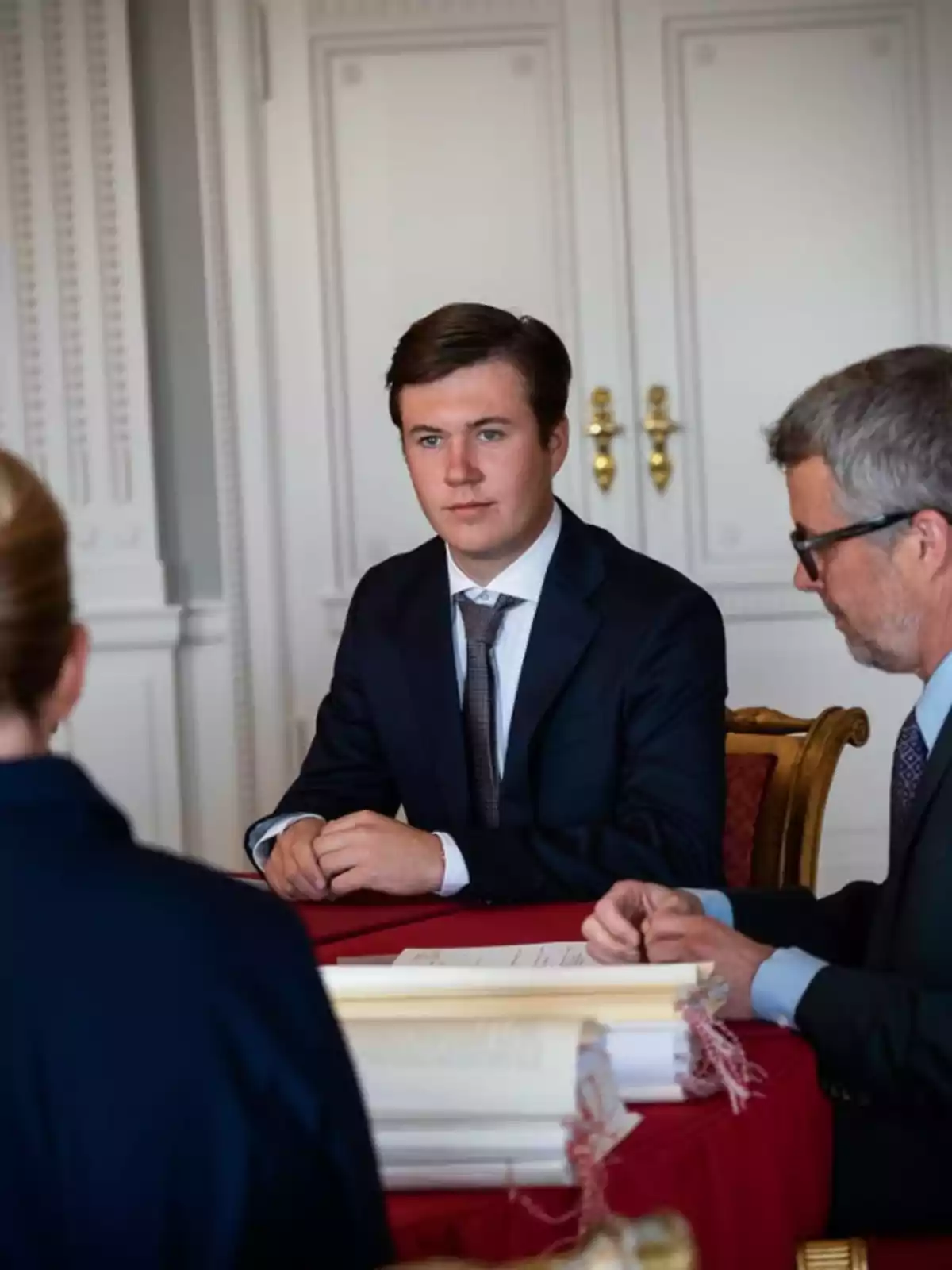 Young man in a dark suit sitting at a red table next to two people in an elegant room Young man in a dark suit sitting at a red table next to two people in an elegant room