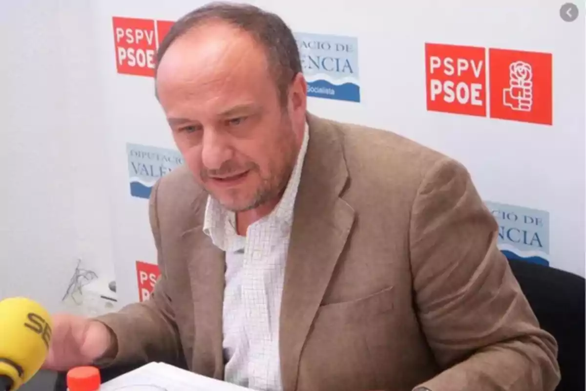 Hombre de mediana edad con chaqueta beige hablando frente a un micrófono amarillo en una rueda de prensa con logotipos de PSPV PSOE y Diputación de Valencia al fondo