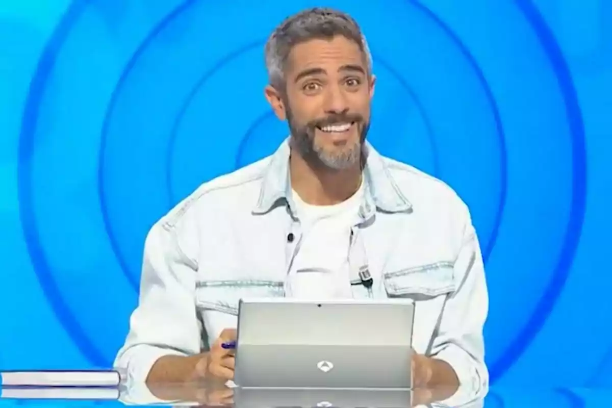 Roberto Leal sonriente con barba y chaqueta clara sentado frente a una tablet en un set de televisión con fondo azul en 'Pasapalabra' Roberto Leal sonriente con barba y chaqueta clara sentado frente a una tablet en un set de televisión con fondo azul en 'Pasapalabra'