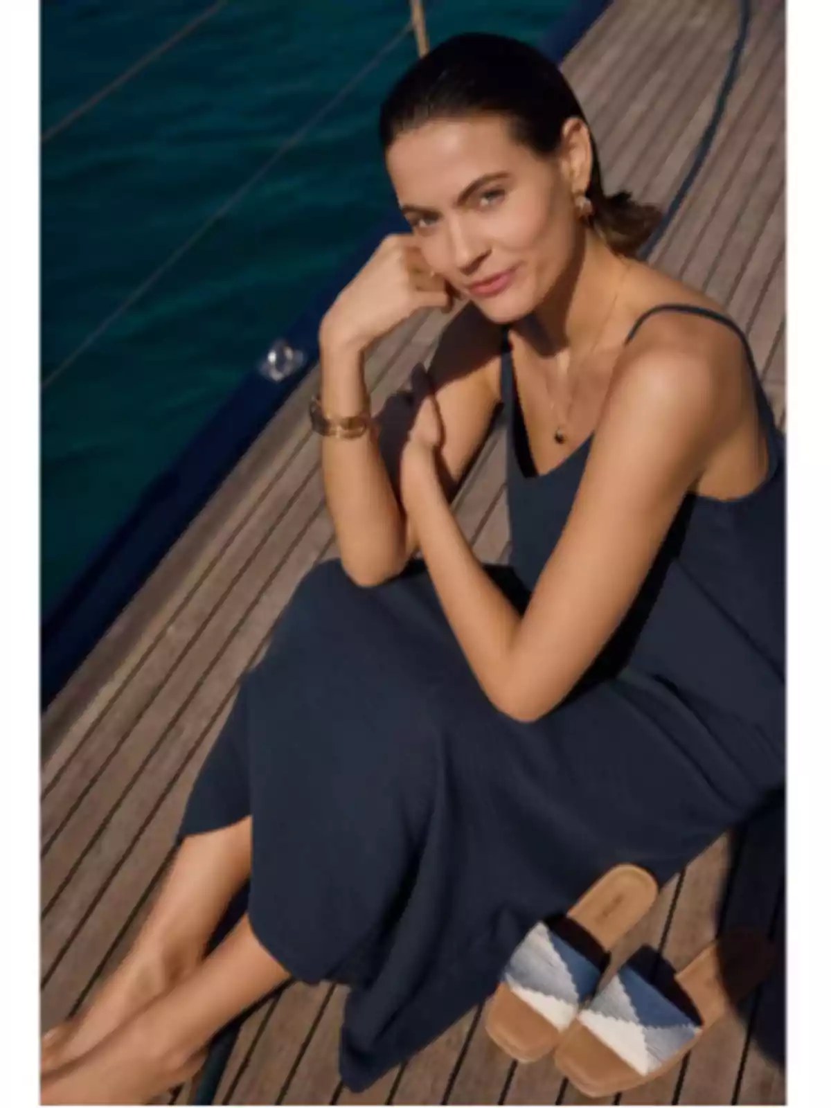 Mujer sentada en la cubierta de un barco con vestido azul marino y sandalias a un lado, mirando a la cámara y sonriendo suavemente Mujer sentada en la cubierta de un barco con vestido azul marino y sandalias a un lado, mirando a la cámara y sonriendo suavemente