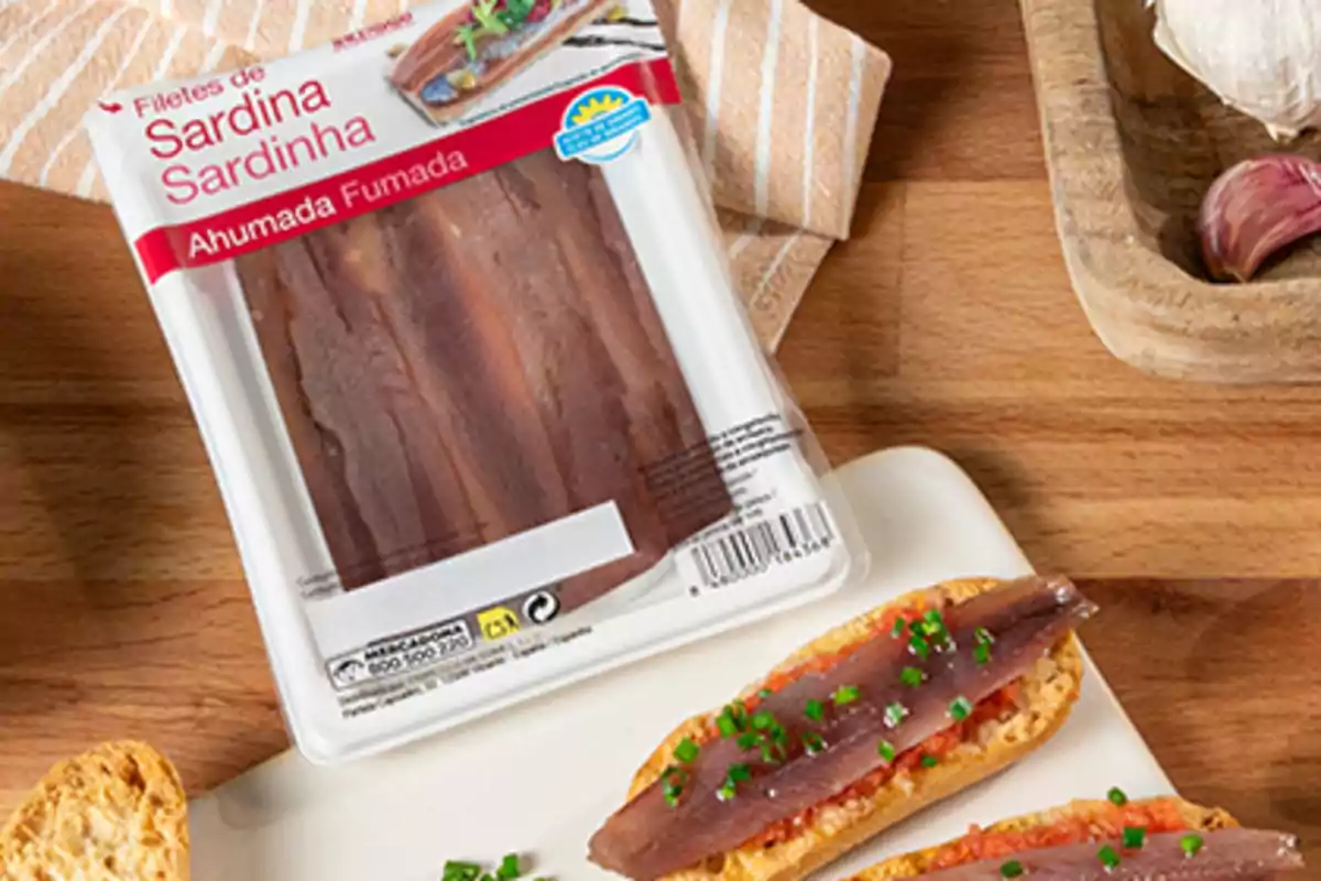 Envase de filetes de sardina ahumada junto a rebanadas de pan con tomate y sardina sobre una mesa de madera Envase de filetes de sardina ahumada junto a rebanadas de pan con tomate y sardina sobre una mesa de madera