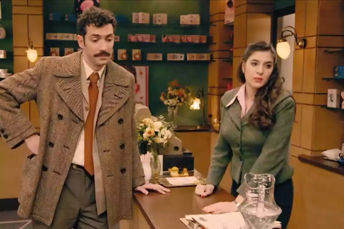 Un hombre con bigote y una mujer de cabello largo están apoyados en un mostrador dentro de una tienda decorada con flores y estanterías llenas de productos. Captura de 'Sueños de libertad' Un hombre con bigote y una mujer de cabello largo están apoyados en un mostrador dentro de una tienda decorada con flores y estanterías llenas de productos. Captura de 'Sueños de libertad'