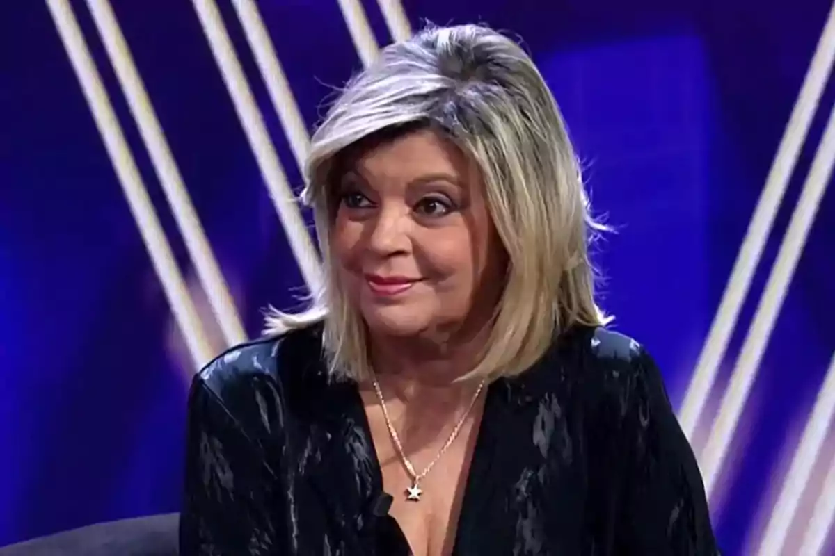 Terelu Campos de cabello rubio y corto con collar de estrella sentada en un set de televisión con fondo azul y líneas blancas Terelu Campos de cabello rubio y corto con collar de estrella sentada en un set de televisión con fondo azul y líneas blancas