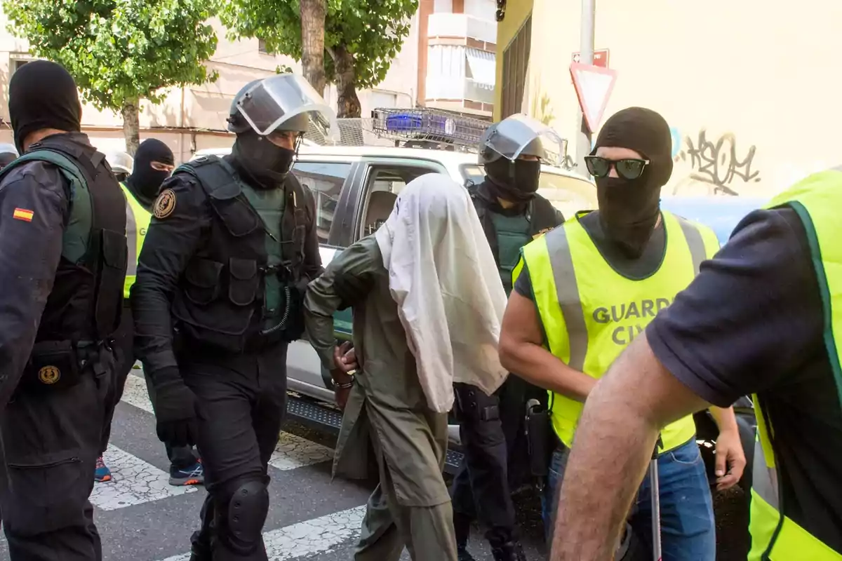 Varios agentes de la Guardia Civil escoltan a una persona detenida con la cabeza cubierta por una tela en una calle urbana Varios agentes de la Guardia Civil escoltan a una persona detenida con la cabeza cubierta por una tela en una calle urbana
