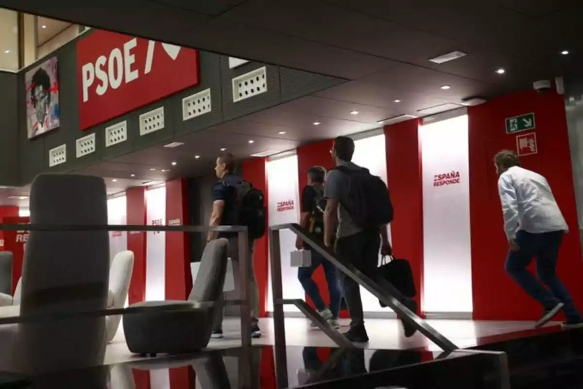 Varias personas suben unas escaleras en el interior de un edificio moderno con paredes rojas y blancas donde se ven los logotipos de PSOE y el lema España Responde. Varias personas suben unas escaleras en el interior de un edificio moderno con paredes rojas y blancas donde se ven los logotipos de PSOE y el lema España Responde.