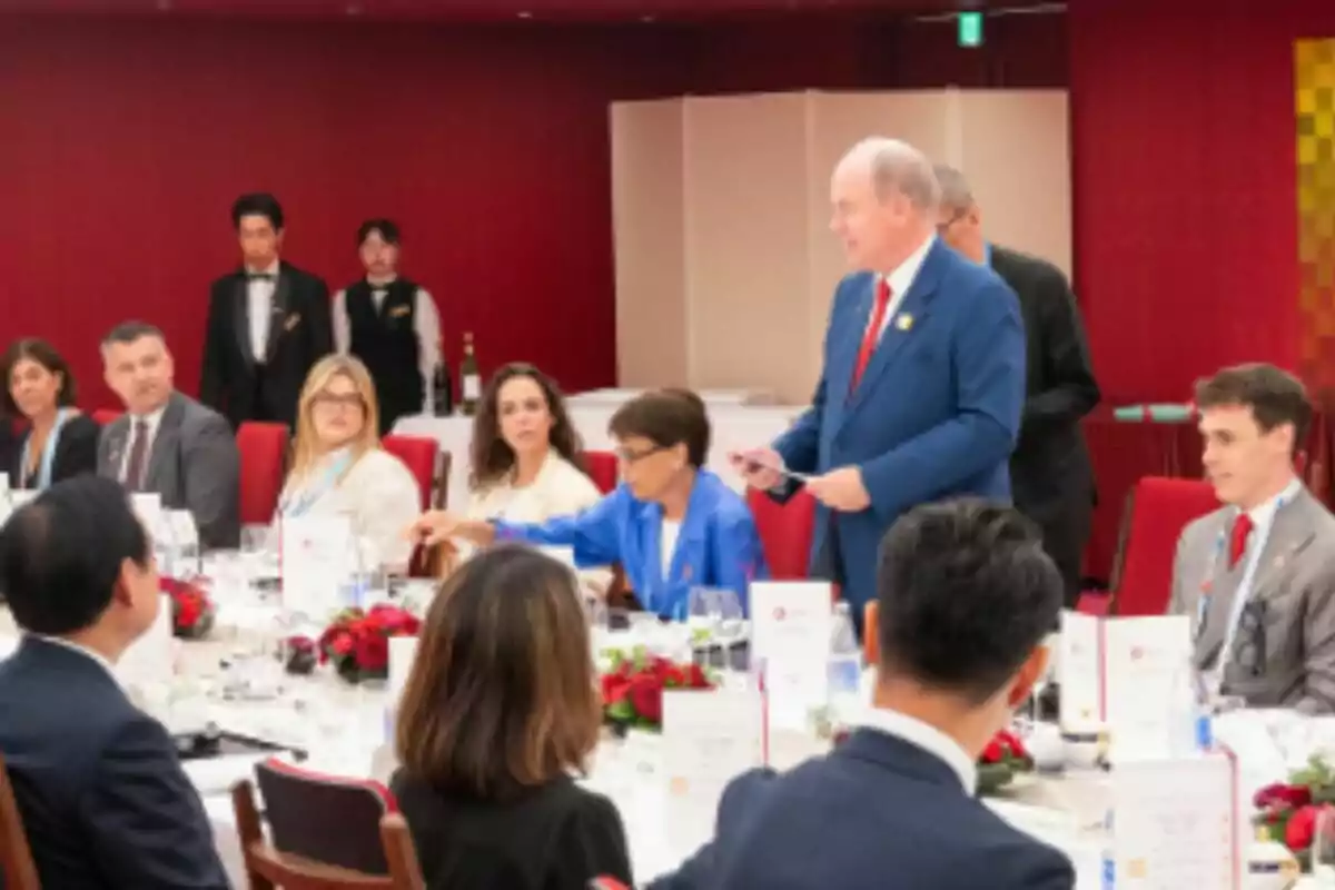 Un grupo de personas elegantemente vestidas participa en una cena formal en un salón decorado en tonos rojos mientras un hombre de traje azul se pone de pie, que es Alberto de Mónaco, y parece estar dando un discurso al lado de Estefanía de Mónaco. Un grupo de personas elegantemente vestidas participa en una cena formal en un salón decorado en tonos rojos mientras un hombre de traje azul se pone de pie, que es Alberto de Mónaco, y parece estar dando un discurso al lado de Estefanía de Mónaco.