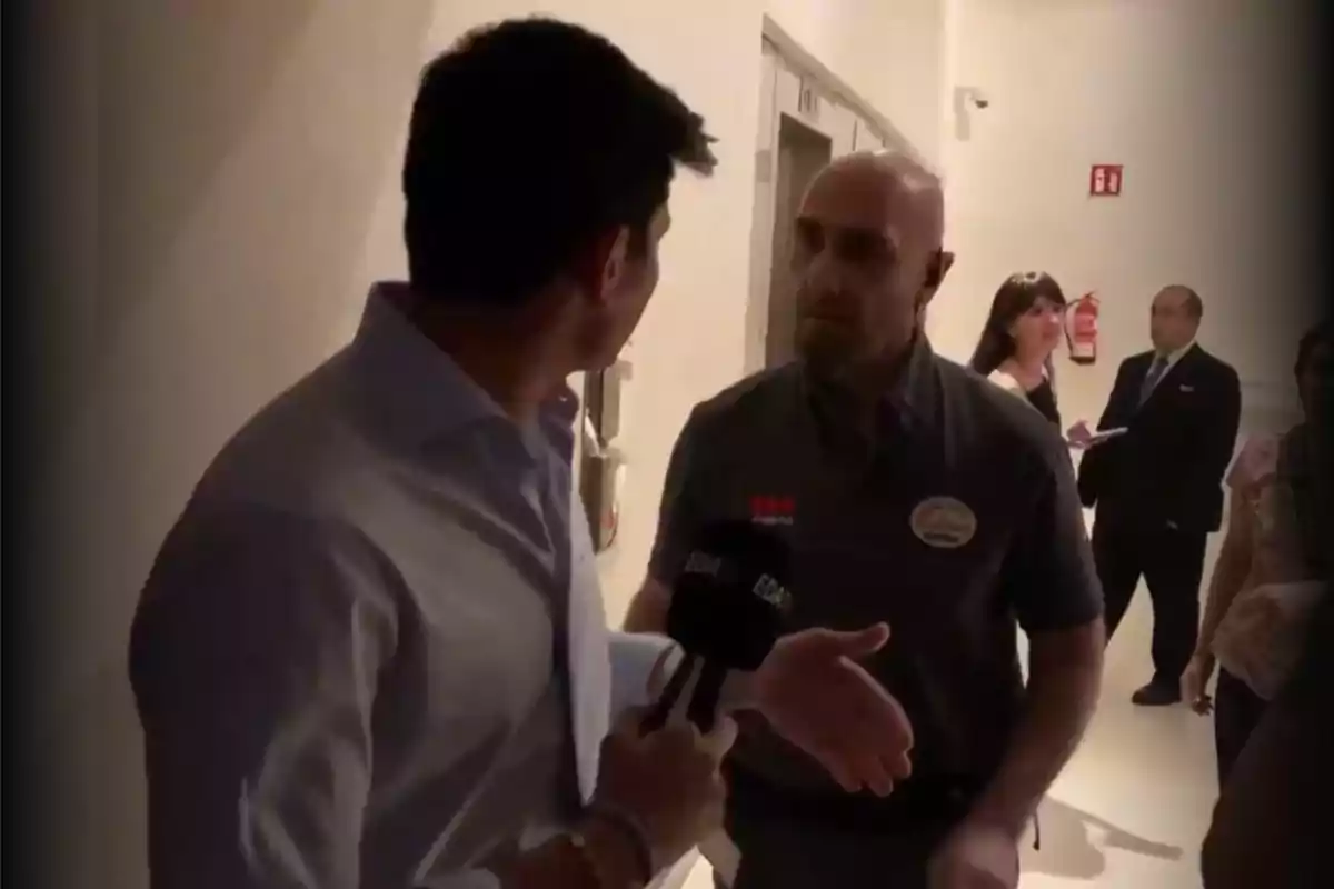 Un reportero sostiene un micrófono mientras conversa con un guardia de seguridad en un pasillo iluminado, al fondo hay más personas y una extintor en la pared.