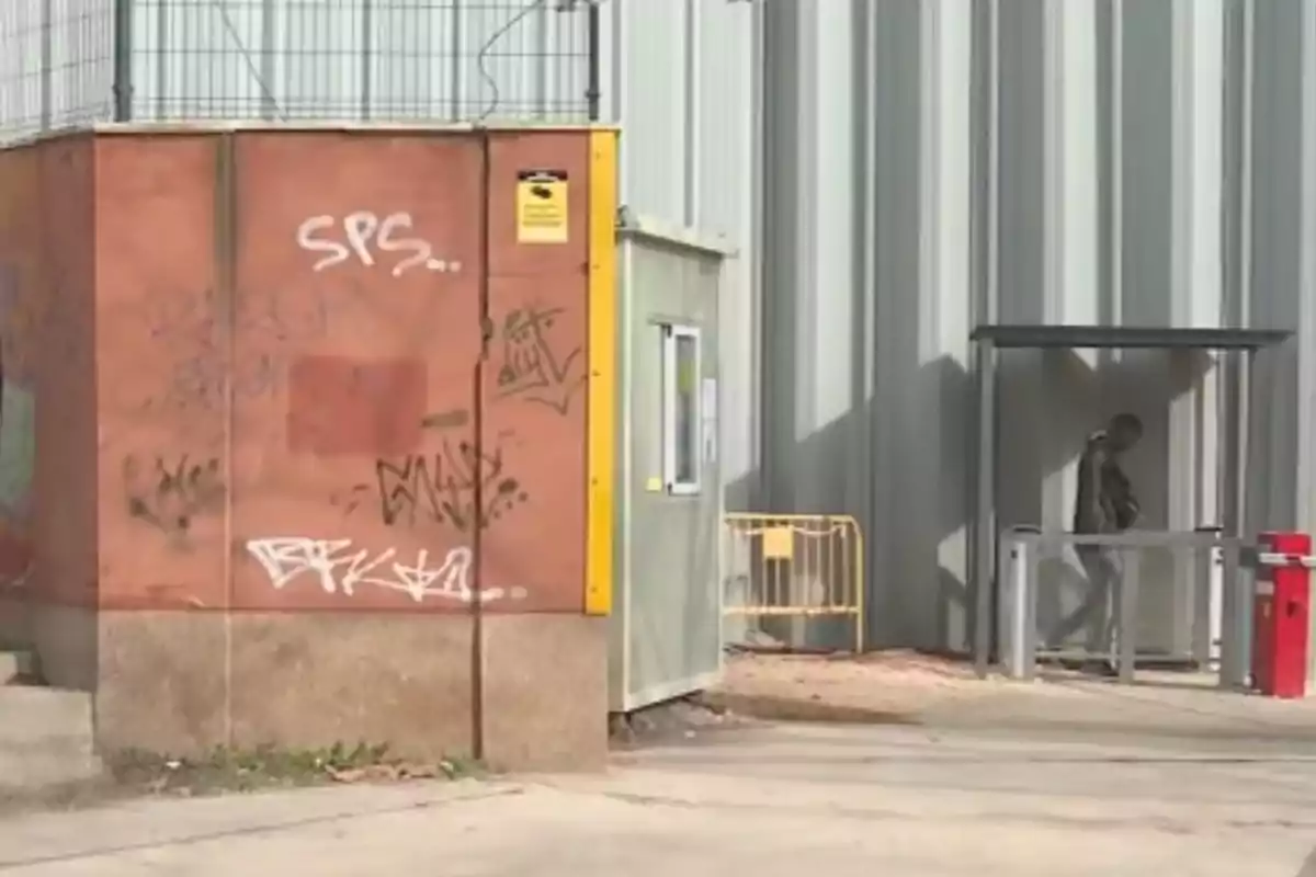 Persona caminando junto a una caseta con grafitis y una estructura metálica industrial al fondo
