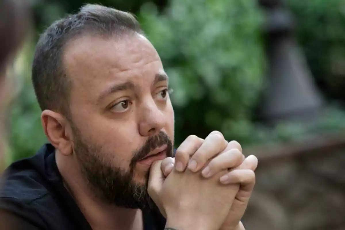 Hombre con barba y cabello corto mirando pensativo mientras apoya las manos entrelazadas en su rostro en un entorno al aire libre con vegetación de fondo
