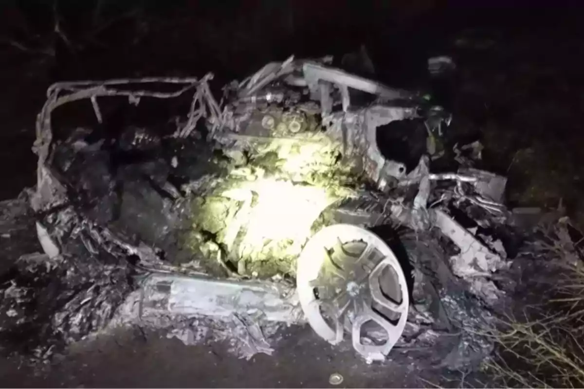 Restos calcinados de un automóvil en la oscuridad con una llanta visible en primer plano y partes metálicas retorcidas iluminadas por una luz fuerte Restos calcinados de un automóvil en la oscuridad con una llanta visible en primer plano y partes metálicas retorcidas iluminadas por una luz fuerte