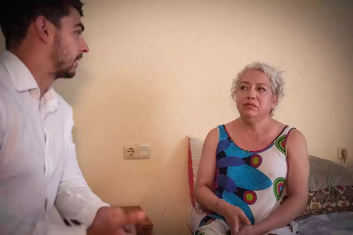 Un hombre y una mujer conversan en una habitación, la mujer está sentada en una cama y parece seria o preocupada.