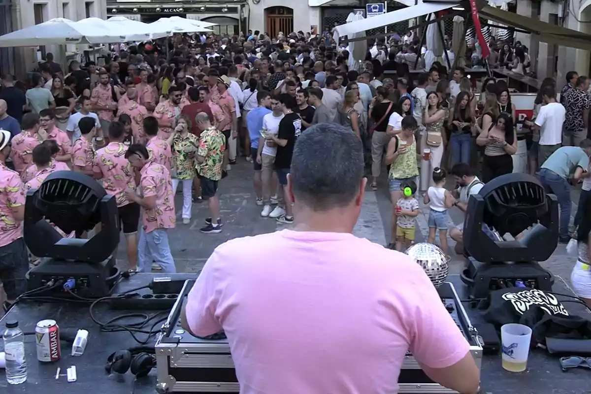 Un DJ de espaldas con camiseta rosa anima una fiesta al aire libre llena de gente, muchos de ellos llevan camisas hawaianas rosas y disfrutan de la música y las bebidas en una calle concurrida Un DJ de espaldas con camiseta rosa anima una fiesta al aire libre llena de gente, muchos de ellos llevan camisas hawaianas rosas y disfrutan de la música y las bebidas en una calle concurrida