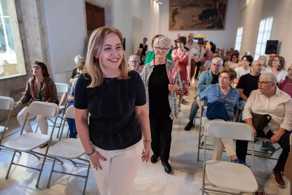 Una mujer sonriente camina entre filas de sillas ocupadas por personas en una sala iluminada, mientras otras personas la siguen y el público observa.