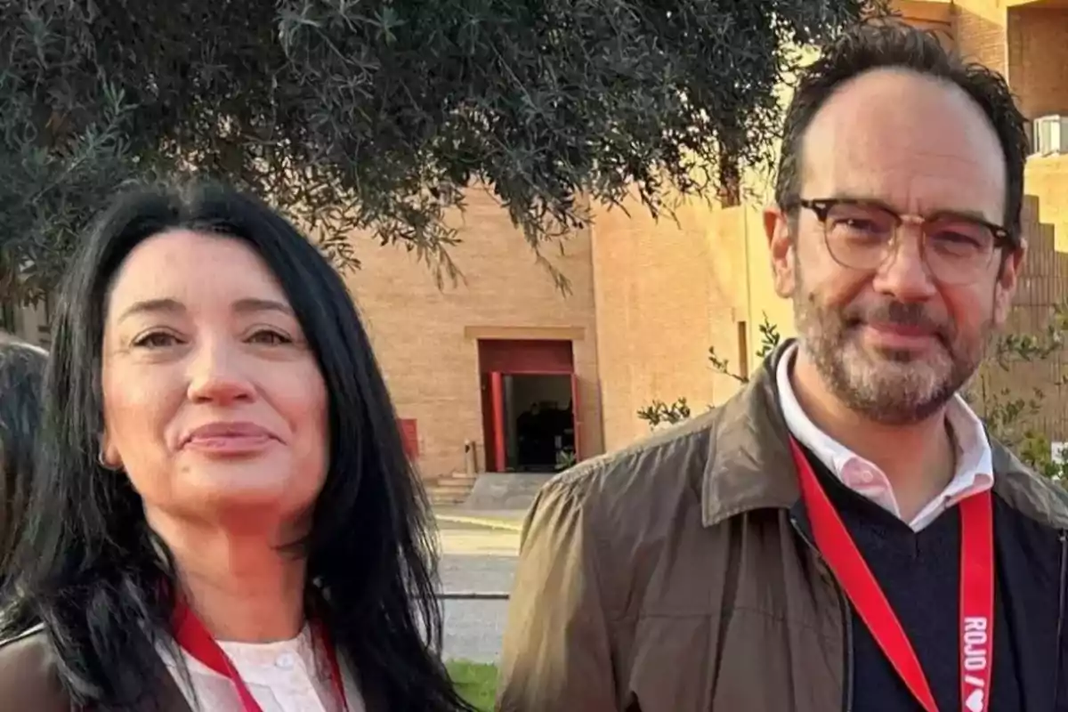 Dos personas posan al aire libre con credenciales rojas colgadas al cuello y un edificio de ladrillo al fondo. Dos personas posan al aire libre con credenciales rojas colgadas al cuello y un edificio de ladrillo al fondo.