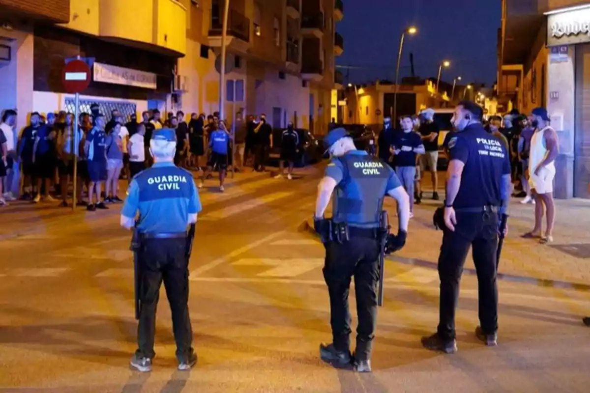 Tres agentes de la Guardia Civil y la Policía Local vigilan una calle por la noche mientras un grupo de personas observa desde la acera Tres agentes de la Guardia Civil y la Policía Local vigilan una calle por la noche mientras un grupo de personas observa desde la acera