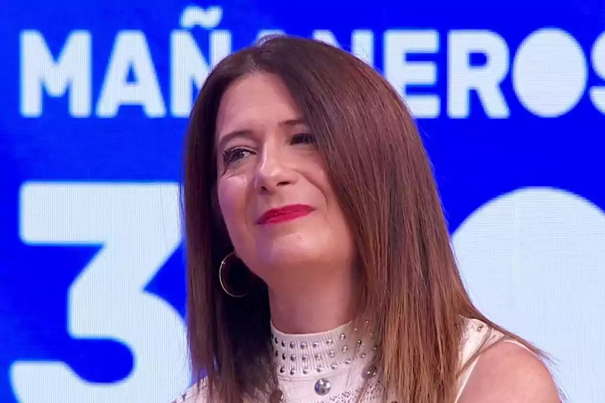 Mujer de cabello lacio castaño y labios rojos sonríe levemente en un set de televisión con fondo azul y letras blancas Mujer de cabello lacio castaño y labios rojos sonríe levemente en un set de televisión con fondo azul y letras blancas