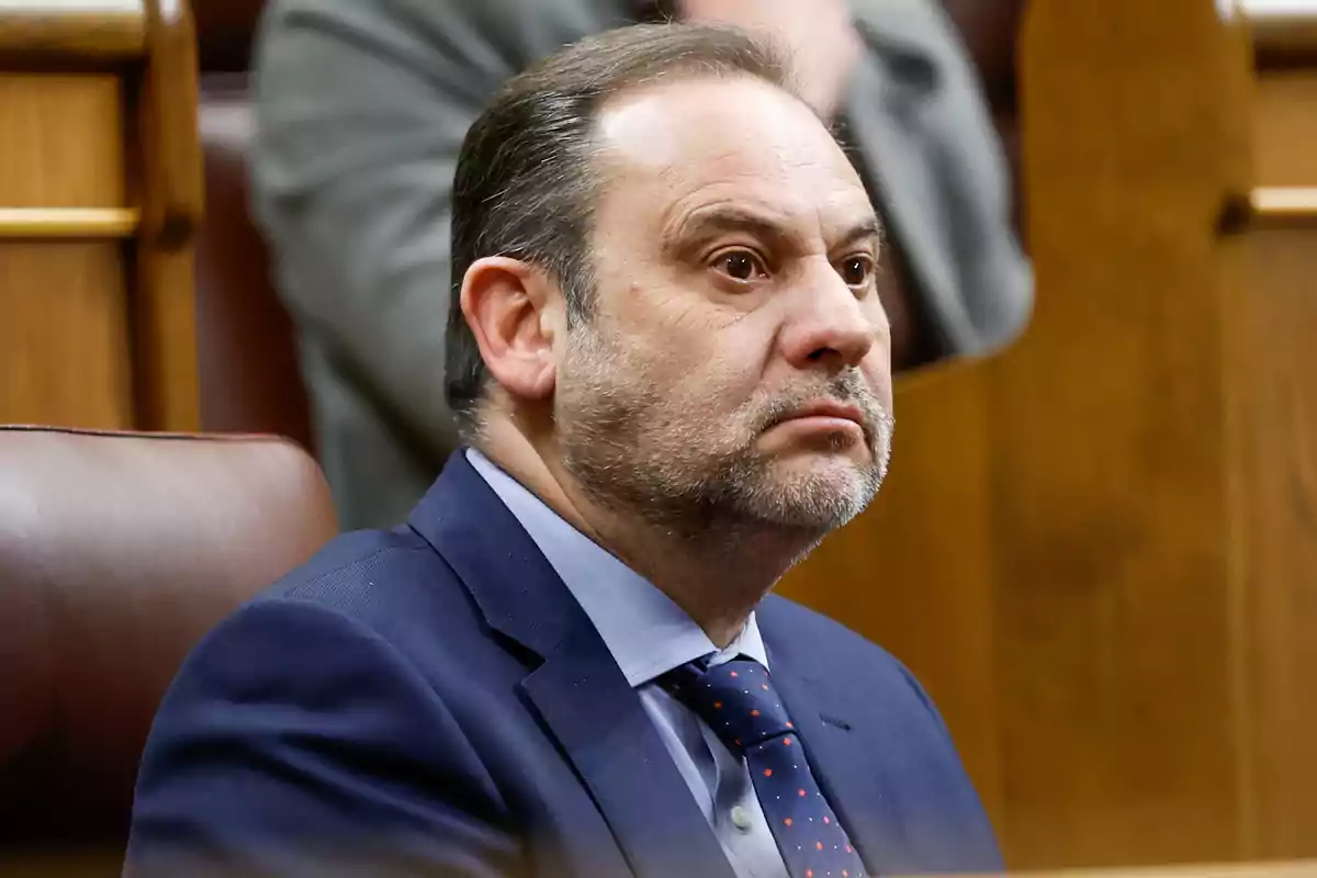 Hombre de mediana edad con barba y traje azul sentado en un escaño de madera en un entorno formal Hombre de mediana edad con barba y traje azul sentado en un escaño de madera en un entorno formal