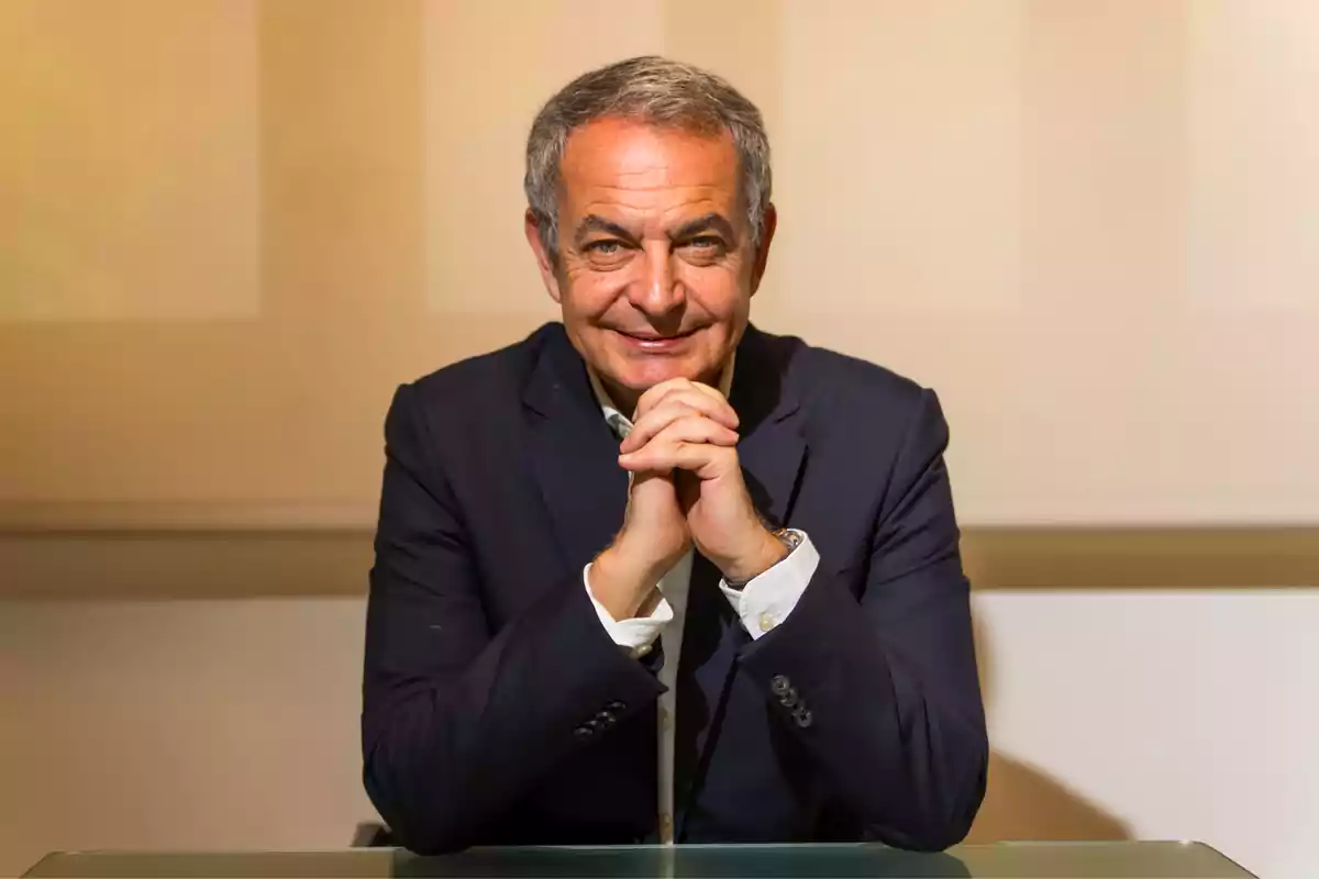 Hombre de cabello canoso con traje azul marino sentado frente a una mesa de cristal con las manos entrelazadas y una expresión sonriente