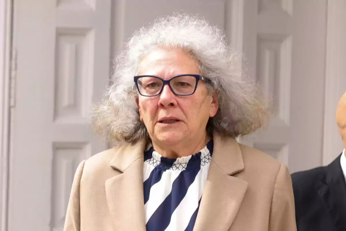 Mujer de cabello canoso y rizado con gafas oscuras, vestida con abrigo beige y blusa de rayas, de pie frente a una puerta blanca Mujer de cabello canoso y rizado con gafas oscuras, vestida con abrigo beige y blusa de rayas, de pie frente a una puerta blanca