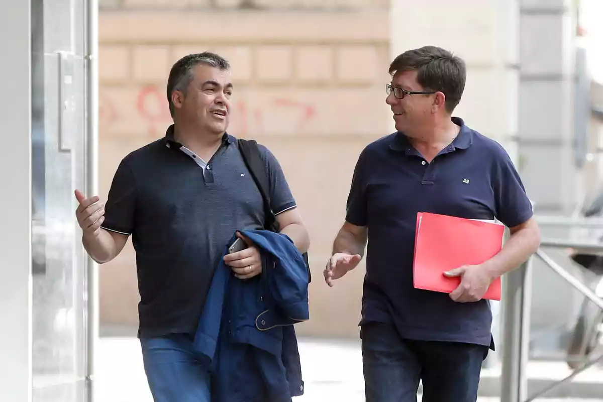 Dos hombres caminan y conversan sonrientes por la calle uno lleva una chaqueta y el otro sostiene una carpeta roja Dos hombres caminan y conversan sonrientes por la calle uno lleva una chaqueta y el otro sostiene una carpeta roja