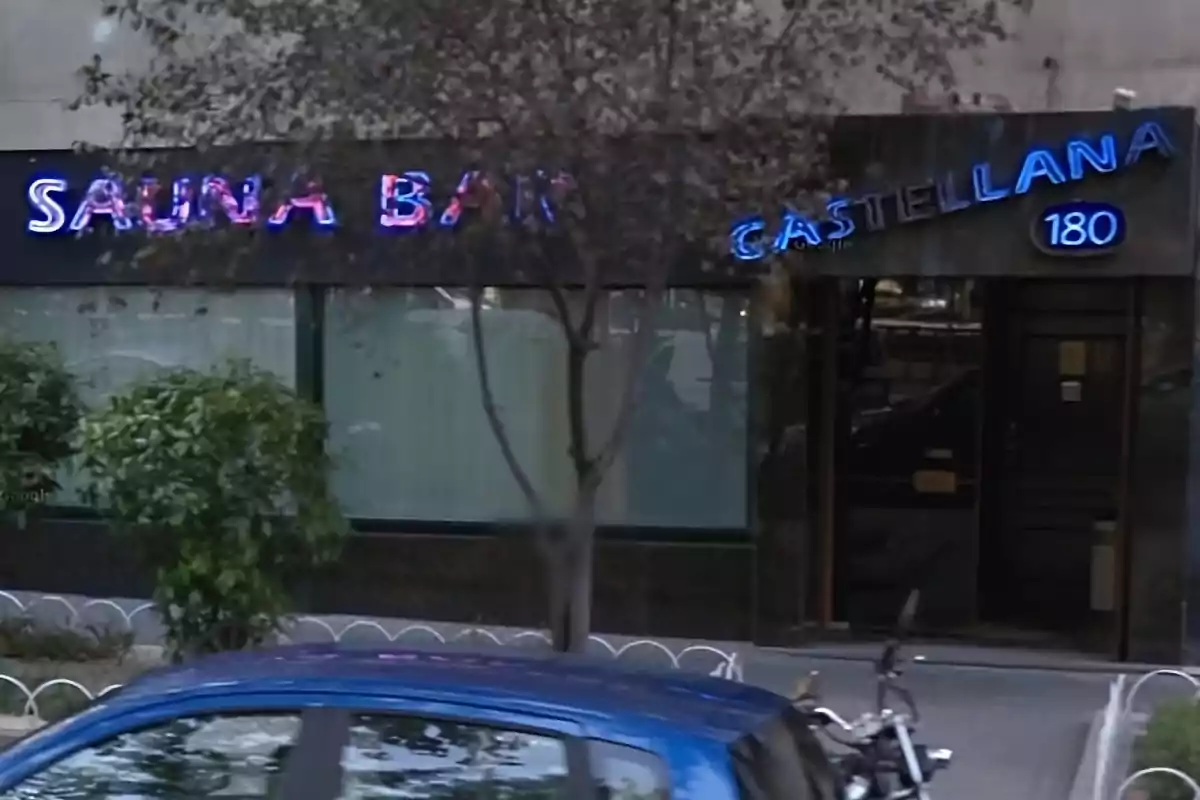 Fachada de un local con letrero de neón que dice Sauna Bar Castellana 180 y un coche azul estacionado al frente Fachada de un local con letrero de neón que dice Sauna Bar Castellana 180 y un coche azul estacionado al frente