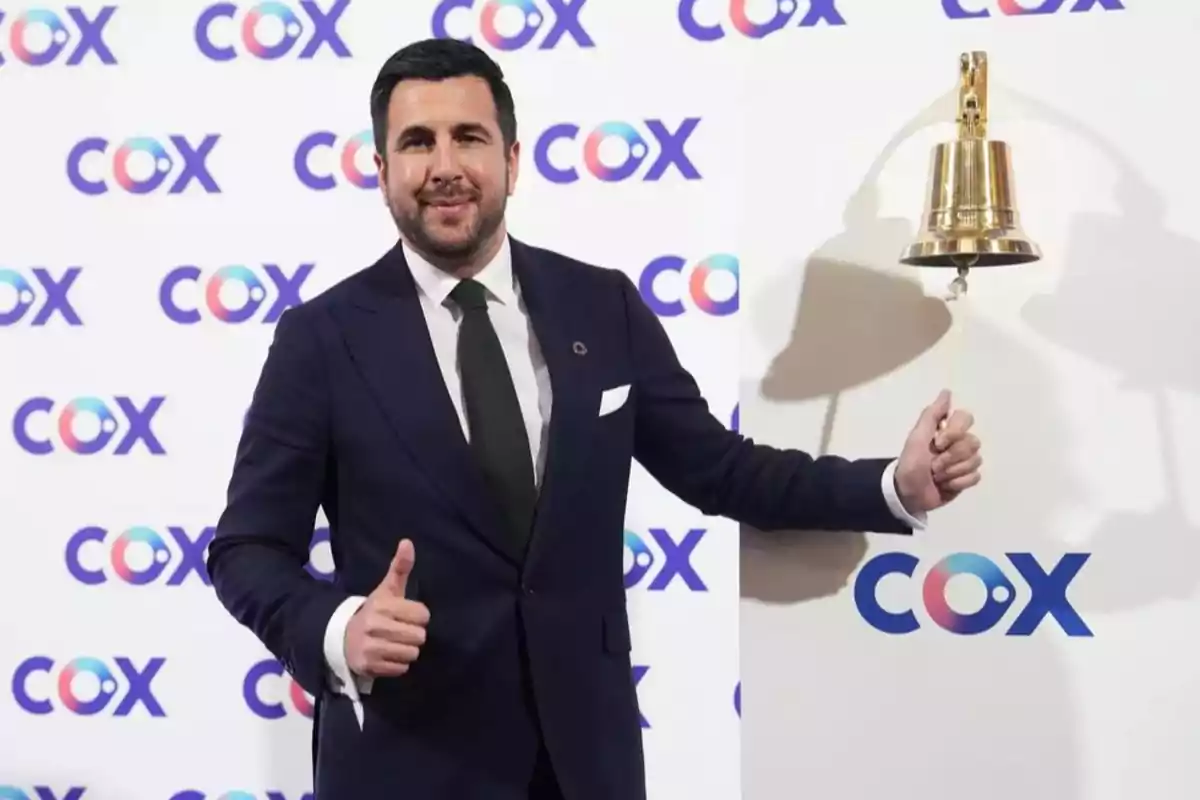 Hombre con traje oscuro tocando una campana dorada y haciendo señal de pulgar arriba frente a un fondo con el logo de COX