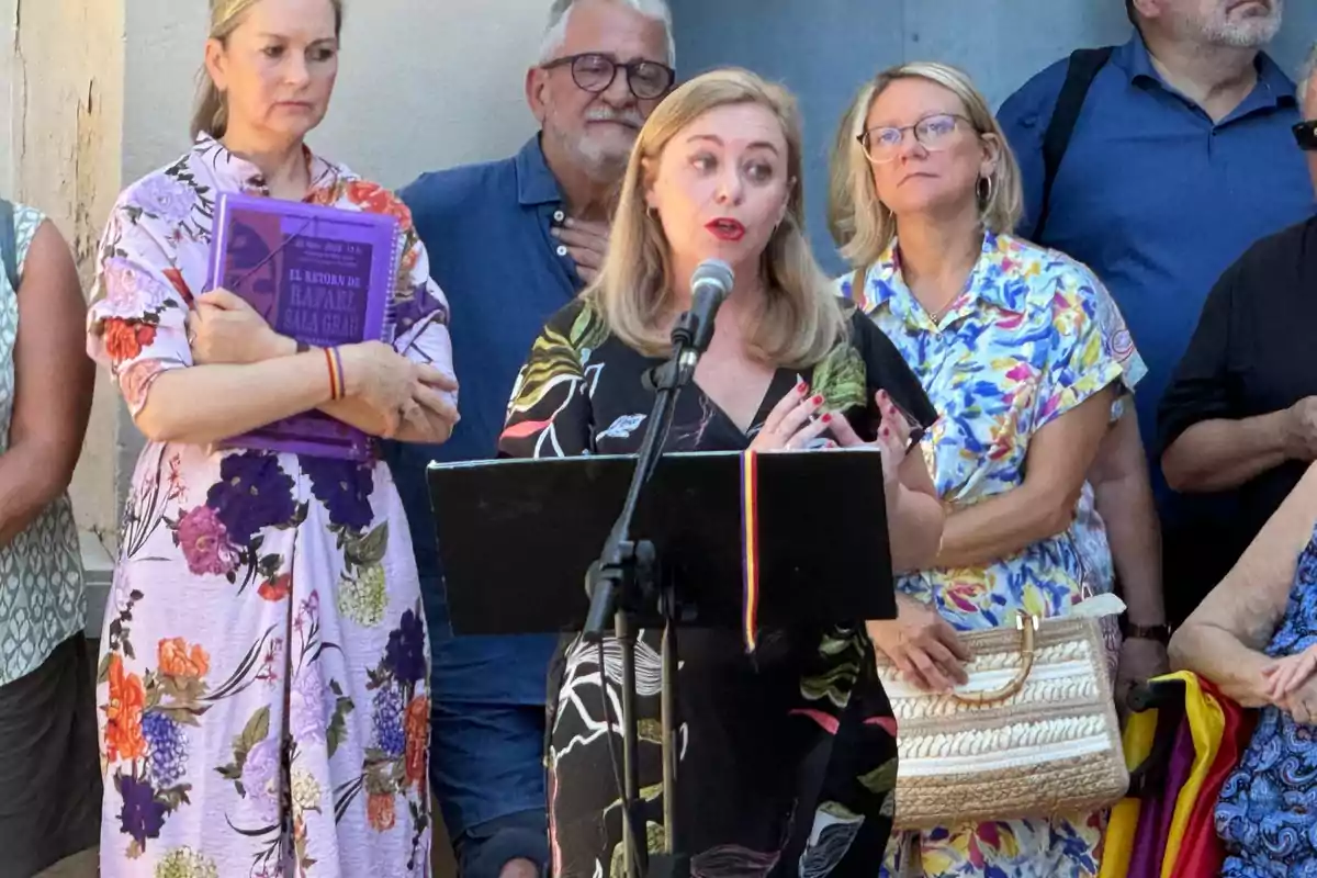 Una mujer rubia habla frente a un micrófono mientras varias personas la escuchan atentamente en un evento al aire libre Una mujer rubia habla frente a un micrófono mientras varias personas la escuchan atentamente en un evento al aire libre