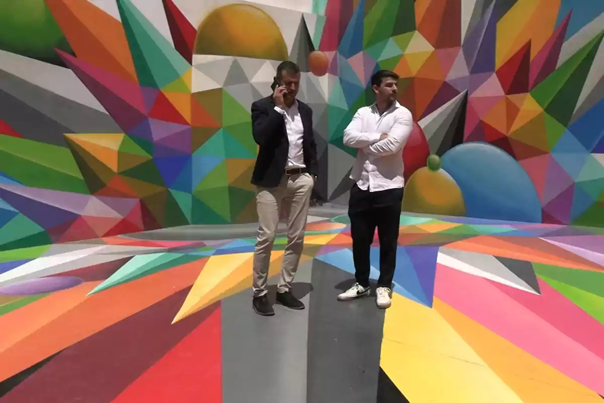 Dos hombres de pie sobre un suelo colorido con un mural geométrico y vibrante de fondo Dos hombres de pie sobre un suelo colorido con un mural geométrico y vibrante de fondo