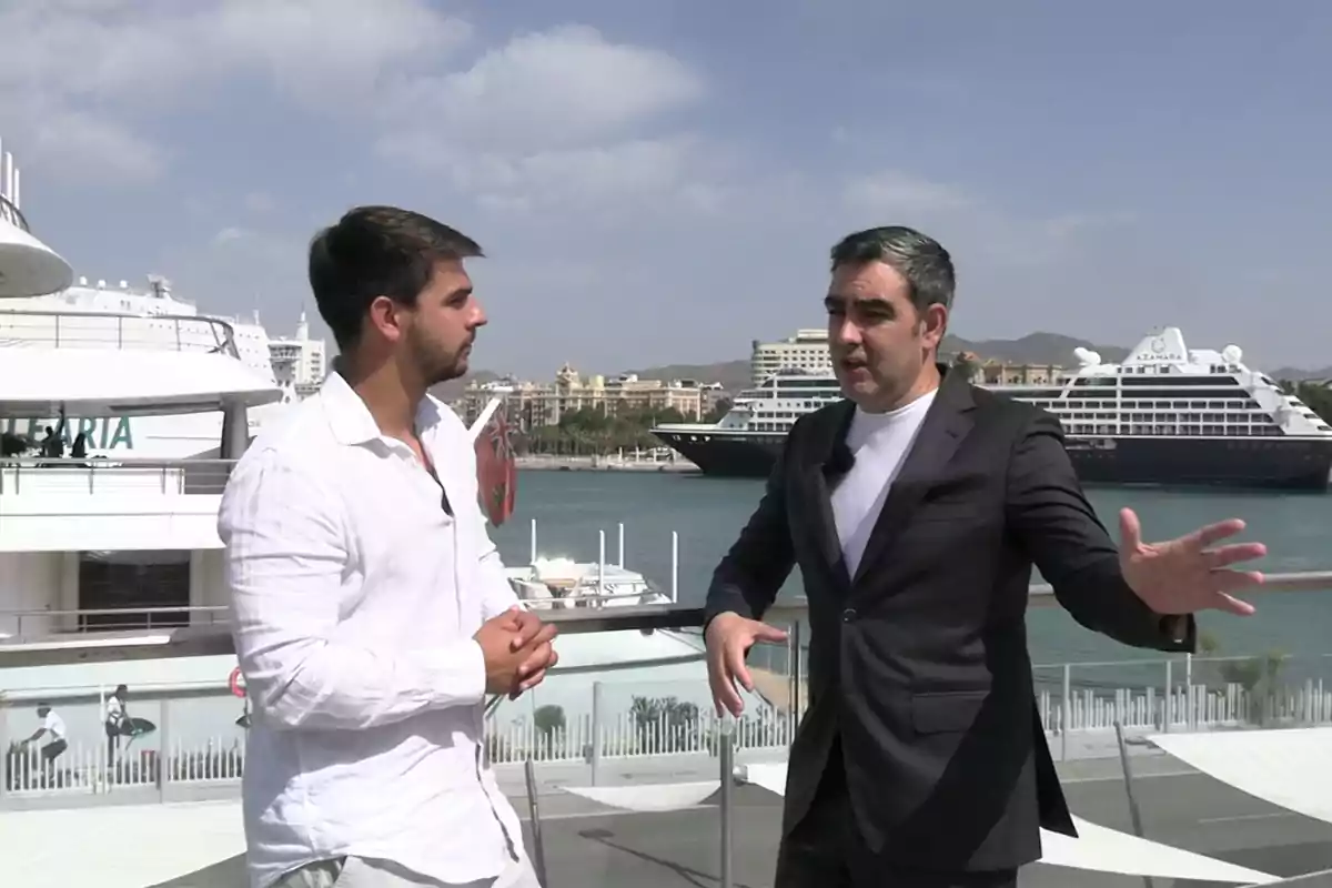 Dos hombres conversan en una terraza con vista al mar y varios cruceros al fondo en un día soleado Dos hombres conversan en una terraza con vista al mar y varios cruceros al fondo en un día soleado