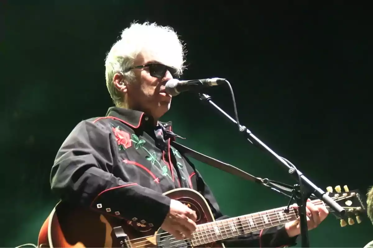 Hombre de cabello canoso y gafas de sol tocando la guitarra y cantando en un escenario con fondo oscuro Hombre de cabello canoso y gafas de sol tocando la guitarra y cantando en un escenario con fondo oscuro