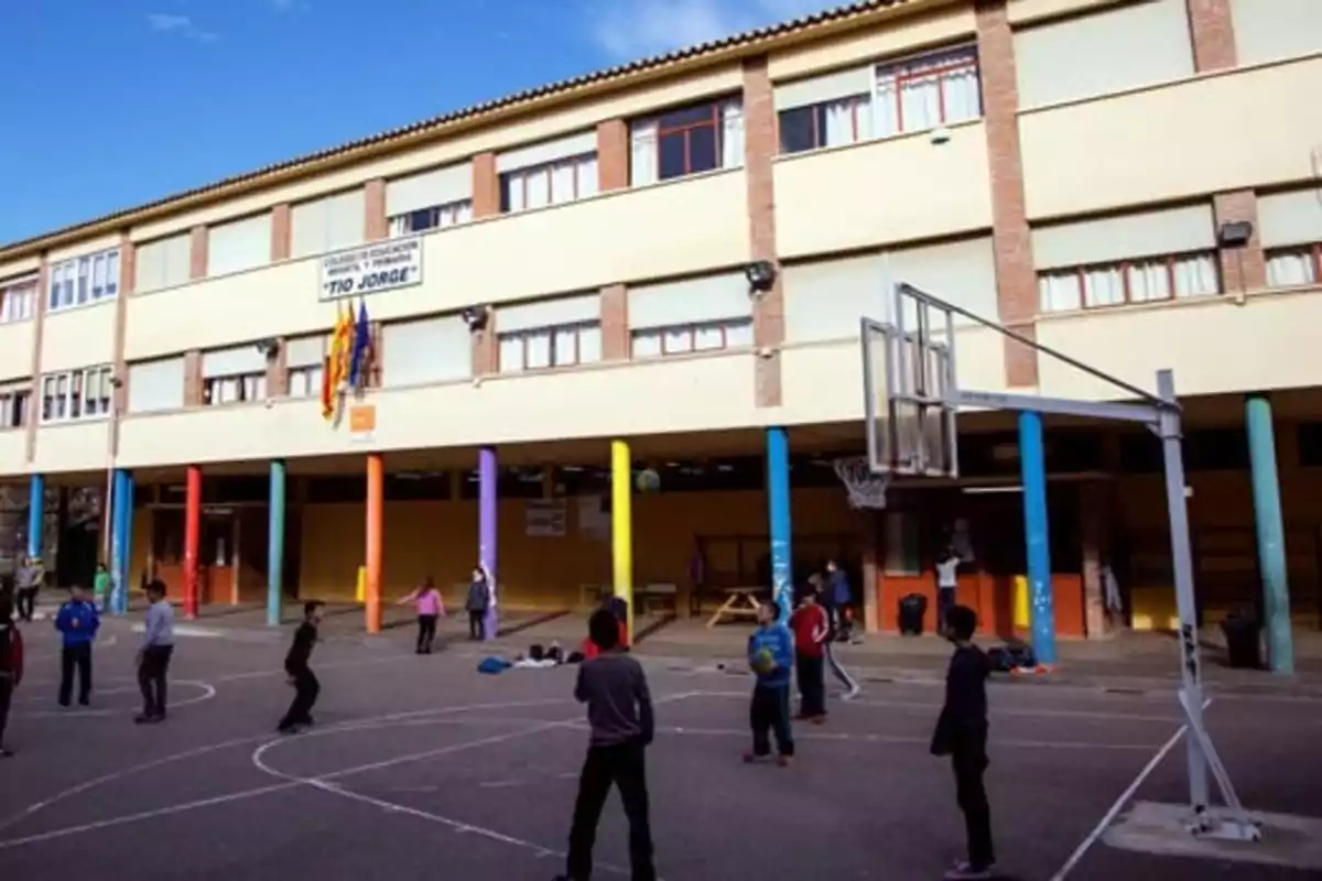 Niños jugando en el patio de una escuela con columnas de colores y una cancha de baloncesto frente a un edificio de varios pisos