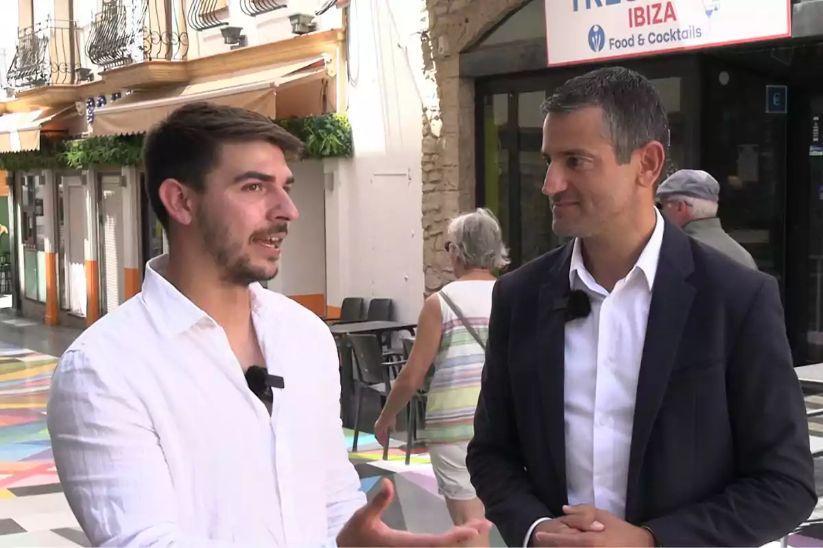 Dos hombres conversan en una calle peatonal colorida frente a un restaurante mientras dos personas caminan al fondo Dos hombres conversan en una calle peatonal colorida frente a un restaurante mientras dos personas caminan al fondo