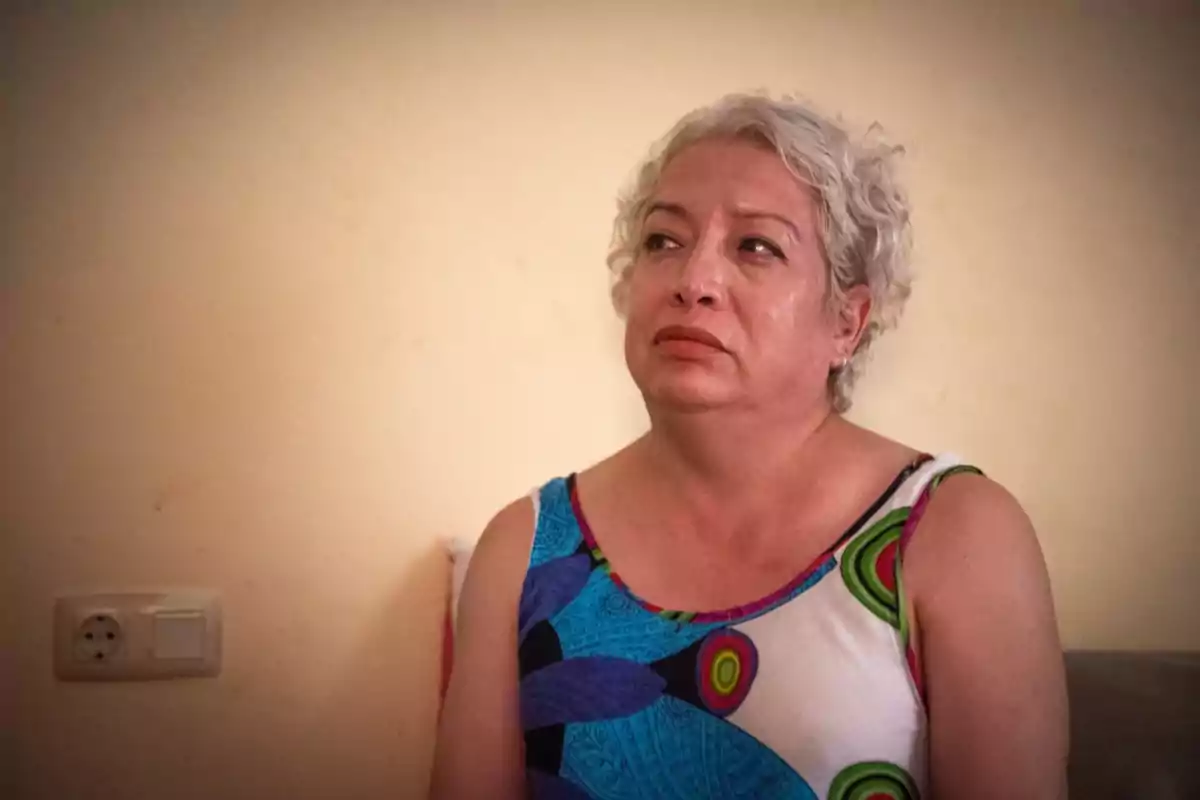 Mujer de cabello corto y canoso con expresión seria, sentada frente a una pared beige y usando un vestido colorido Mujer de cabello corto y canoso con expresión seria, sentada frente a una pared beige y usando un vestido colorido