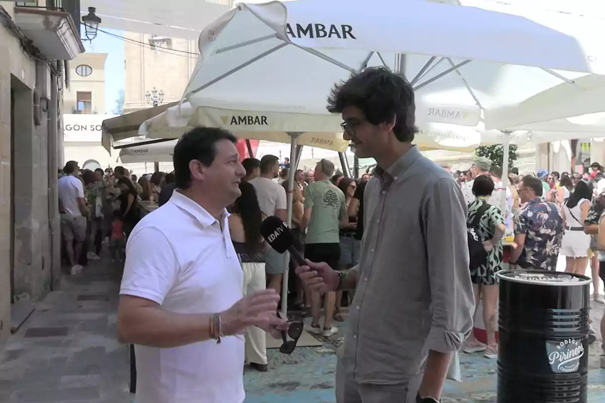 Un hombre con camisa blanca es entrevistado por un reportero con camisa gris en una calle concurrida durante un evento al aire libre con sombrillas y muchas personas al fondo Un hombre con camisa blanca es entrevistado por un reportero con camisa gris en una calle concurrida durante un evento al aire libre con sombrillas y muchas personas al fondo