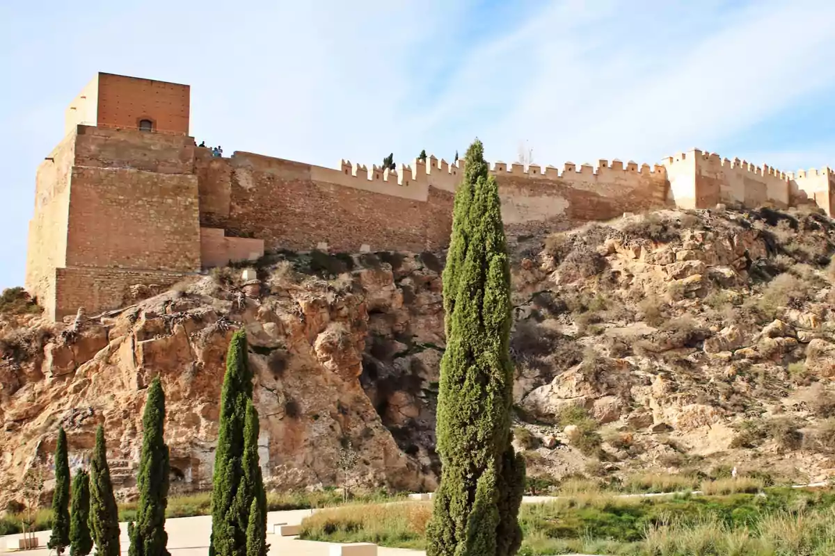Murallas de una fortaleza antigua construida sobre una colina rocosa con cipreses en primer plano y cielo despejado