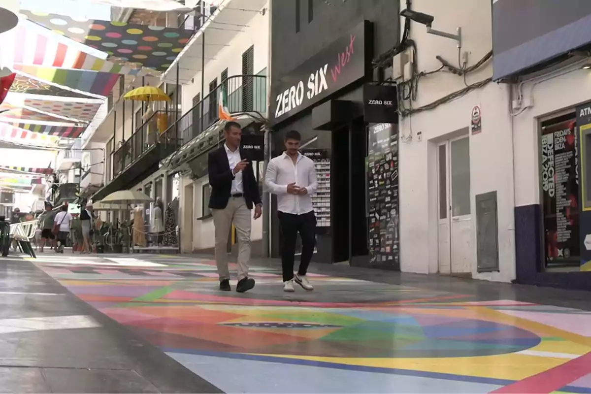 Dos hombres caminan por una calle peatonal con el suelo pintado de colores y toldos decorativos en la parte superior, rodeados de tiendas y personas al fondo Dos hombres caminan por una calle peatonal con el suelo pintado de colores y toldos decorativos en la parte superior, rodeados de tiendas y personas al fondo