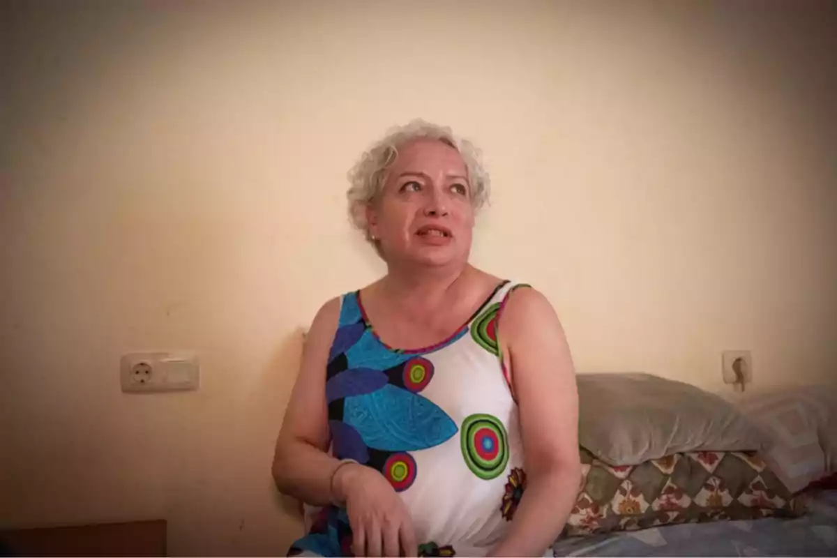 Mujer de cabello corto y canoso sentada en una cama con cojines, lleva un vestido colorido y mira hacia un lado en una habitación de paredes claras Mujer de cabello corto y canoso sentada en una cama con cojines, lleva un vestido colorido y mira hacia un lado en una habitación de paredes claras