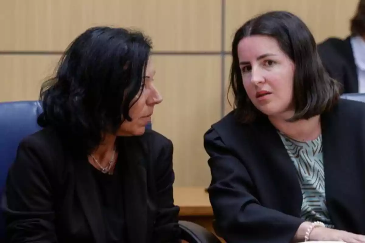 Dos mujeres sentadas conversan en lo que parece ser una sala de reuniones o tribunal, ambas visten ropa formal y muestran expresiones serias. Dos mujeres sentadas conversan en lo que parece ser una sala de reuniones o tribunal, ambas visten ropa formal y muestran expresiones serias.