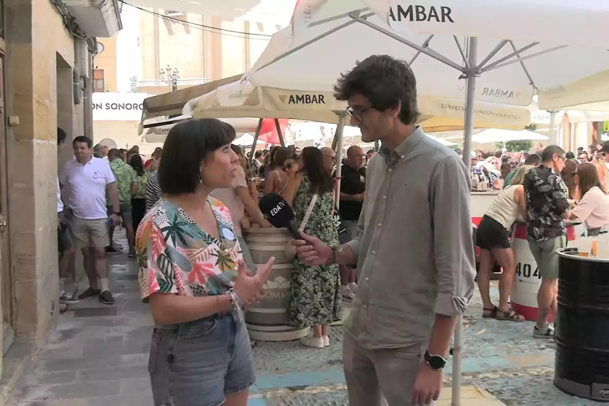 Un hombre joven entrevista a una mujer en una terraza concurrida durante un evento al aire libre, rodeados de personas y sombrillas de un bar Un hombre joven entrevista a una mujer en una terraza concurrida durante un evento al aire libre, rodeados de personas y sombrillas de un bar