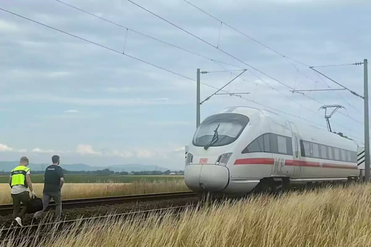 Dos personas con chalecos de policía caminan junto a las vías donde está detenido un tren de alta velocidad en un entorno rural Dos personas con chalecos de policía caminan junto a las vías donde está detenido un tren de alta velocidad en un entorno rural