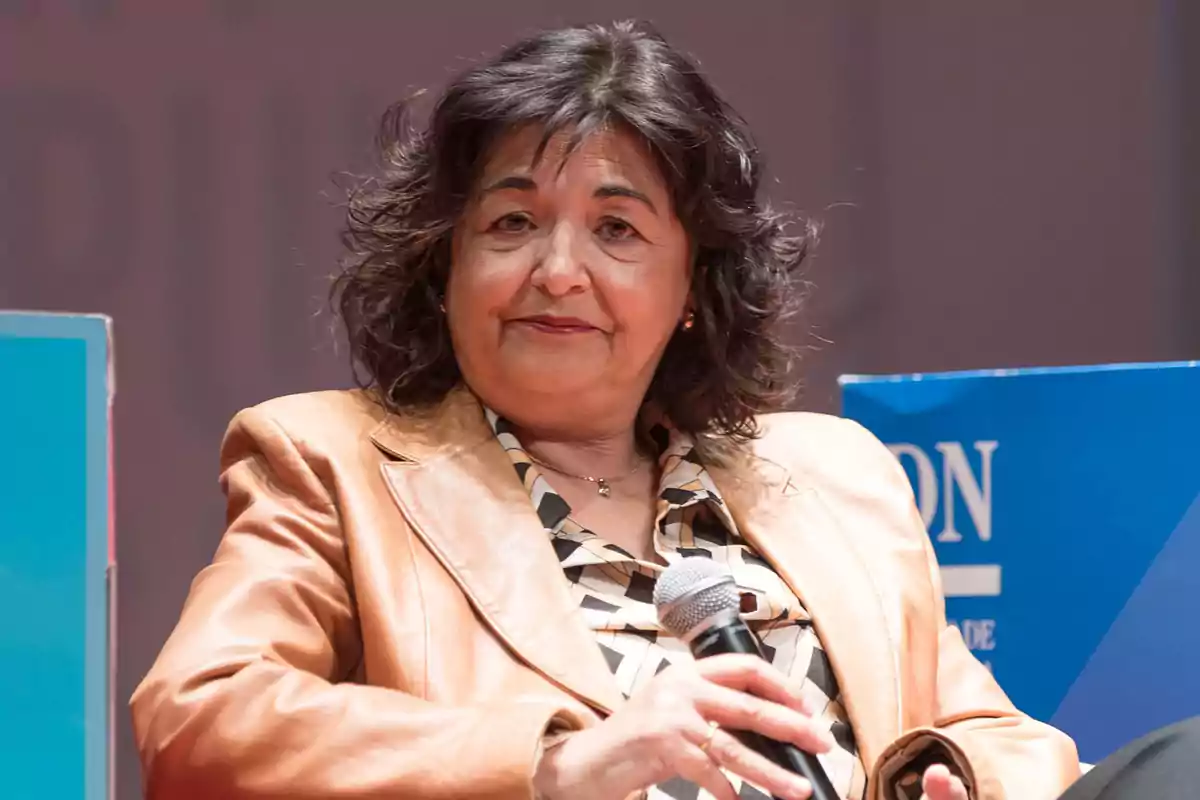 Mujer de cabello oscuro y ondulado con chaqueta beige sosteniendo un micrófono durante un evento Mujer de cabello oscuro y ondulado con chaqueta beige sosteniendo un micrófono durante un evento